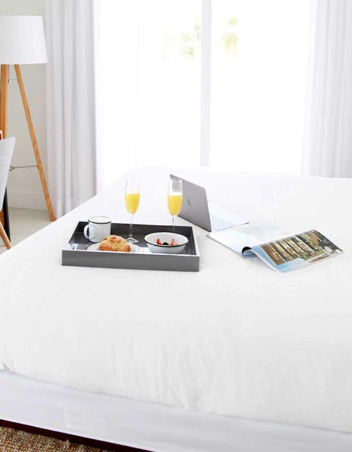 Breakfast tray with food, drinks, laptop, and magazine on a white bed near a window.