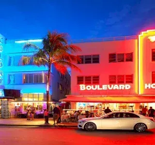 Night scene of a Miami street with neon-lit art deco buildings, palm trees, and a parked car.