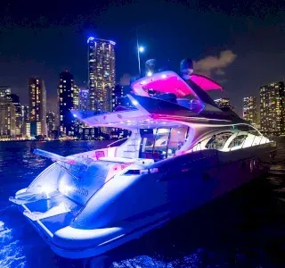 Luxury yacht illuminated with blue and pink lights at night, skyline in the background.