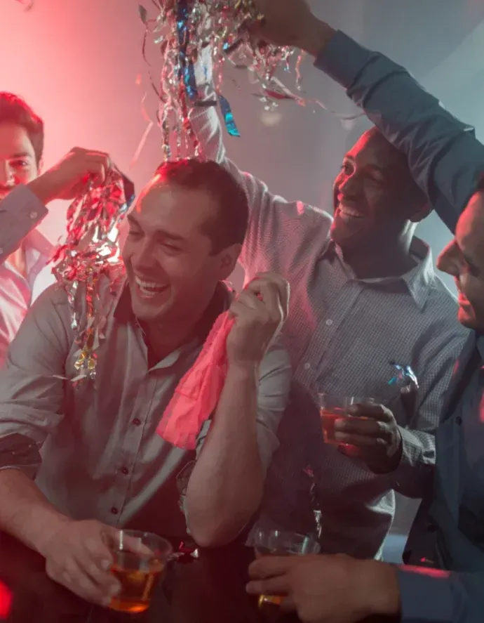 Men laughing, celebrating at a bar with drinks and streamers, lit with red and blue.