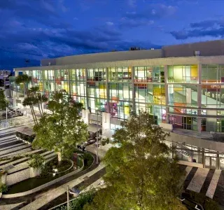 Modern building with large windows illuminated at dusk, colorful interior. Trees and paved walkway in foreground.