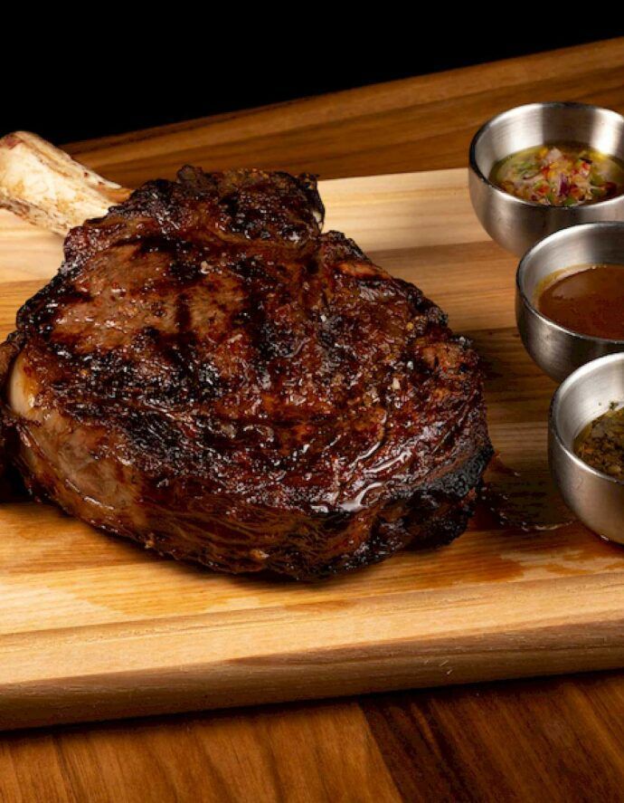 Grilled tomahawk steak on a wooden board with small bowls of sauce.