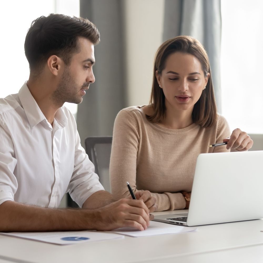 A man and a woman are sitting at a table looking at a laptop computer.