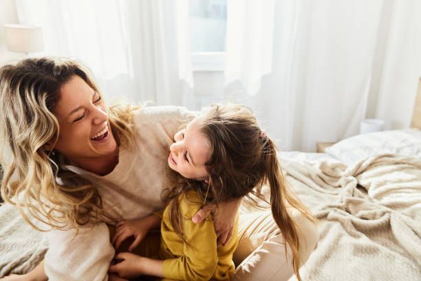 Woman and child laughing on a bed; woman in light colors, child in yellow; natural light.