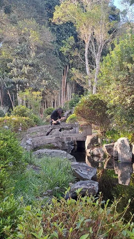 Mateus Asato tocando la guitarra en un puente de estilo japonés rodeado de vegetación.