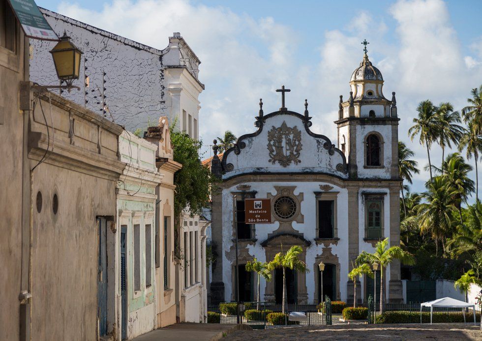 A church with a clock tower is surrounded by buildings and palm trees.