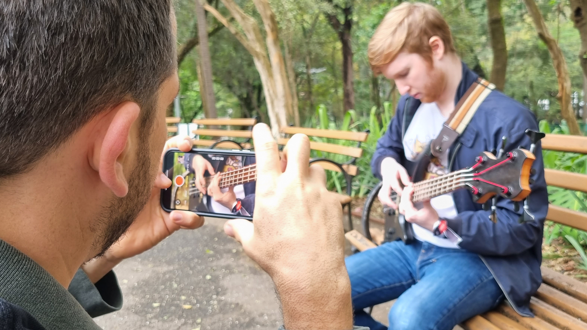A man is taking a picture of a man playing a guitar on a bench.
