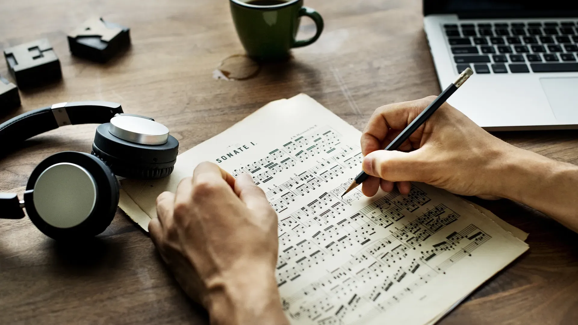 A person is writing on a piece of music next to a laptop and headphones.