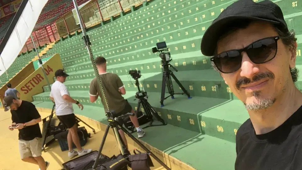 A man wearing sunglasses and a hat is standing in front of a stadium.