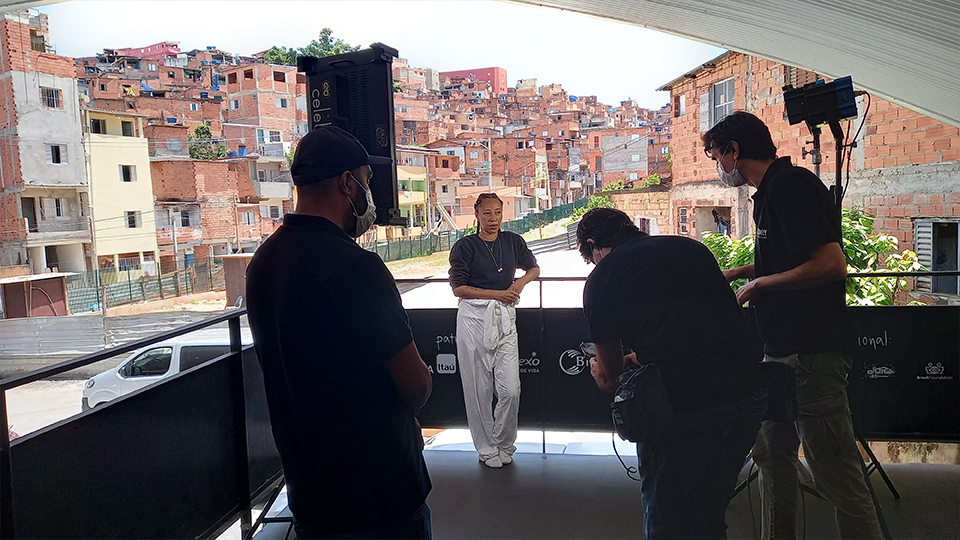 A woman stands in front of a crew filming with the slums as a background