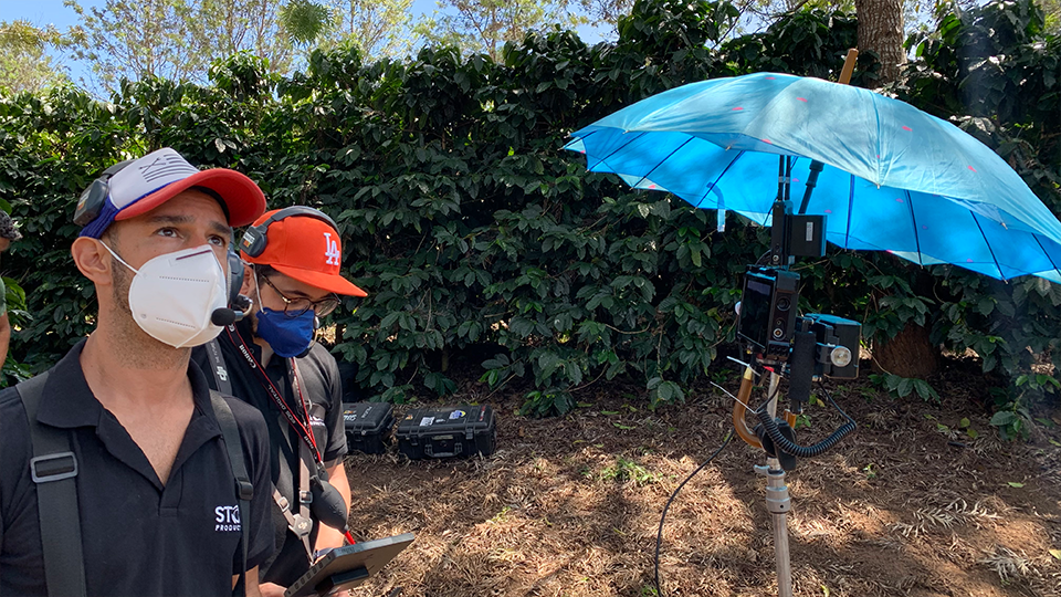 Story Productions crew films on location A man wearing a mask and a hat is standing next to a blue umbrella.