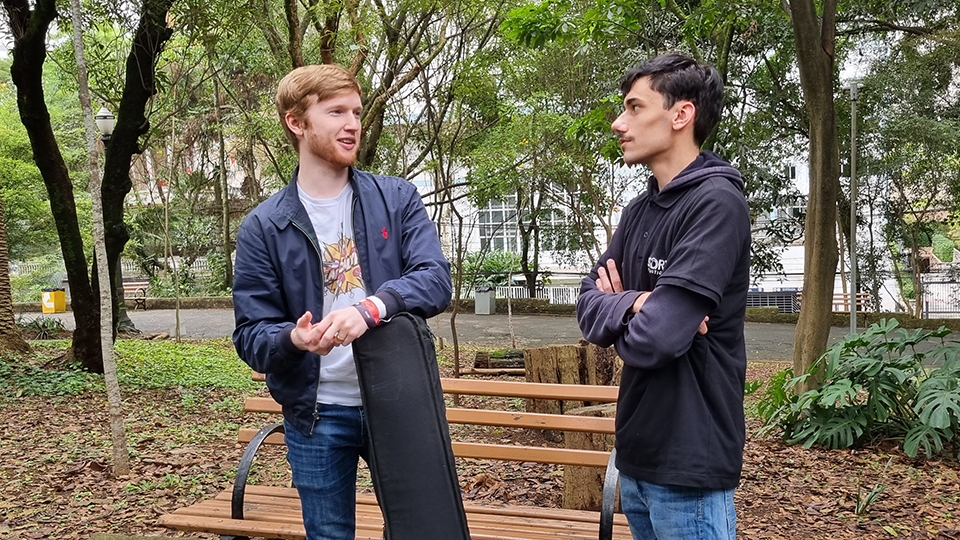 Two men are standing next to each other in a park holding a skateboard.