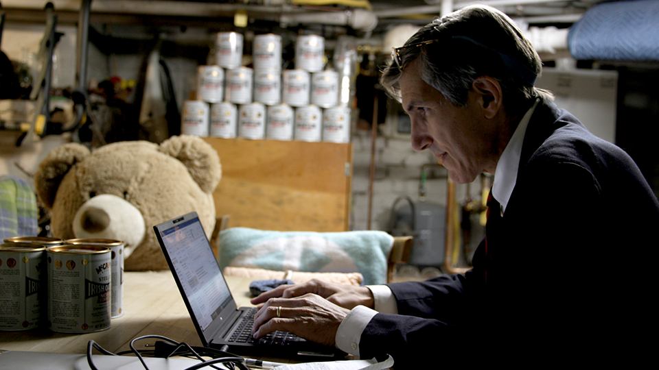 A man is typing on a laptop in a garage with a teddy bear in the background.