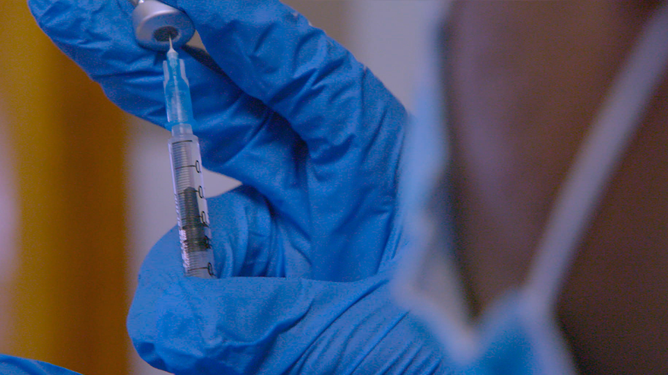 A person wearing blue gloves is holding a syringe with a vaccine in it.
