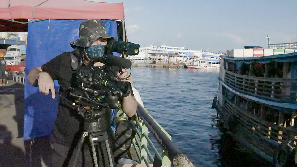 Story Productions films on a location in Manaus A man is standing on a boat holding a camera.