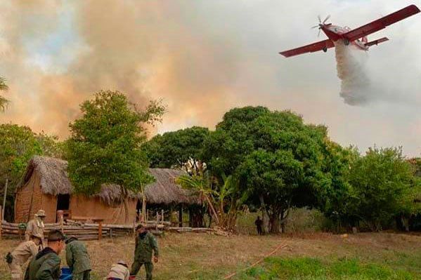A plane flies above a burning field with a wooden shack