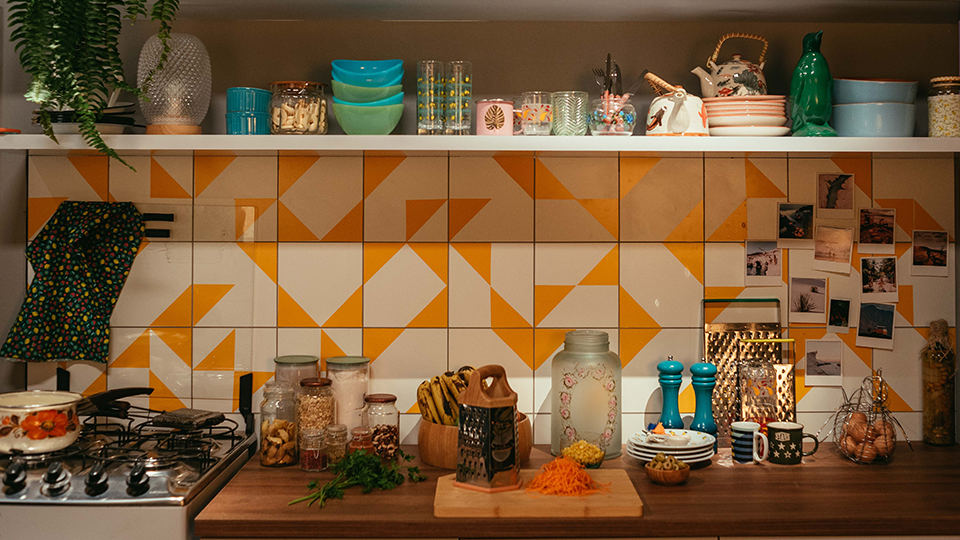 A kitchen with yellow and white tiles , a cutting board , a stove and a shelf.