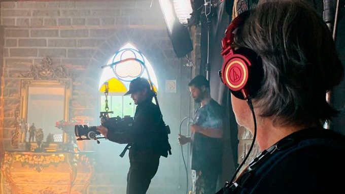 A man is holding a camera while a woman wearing headphones watches.