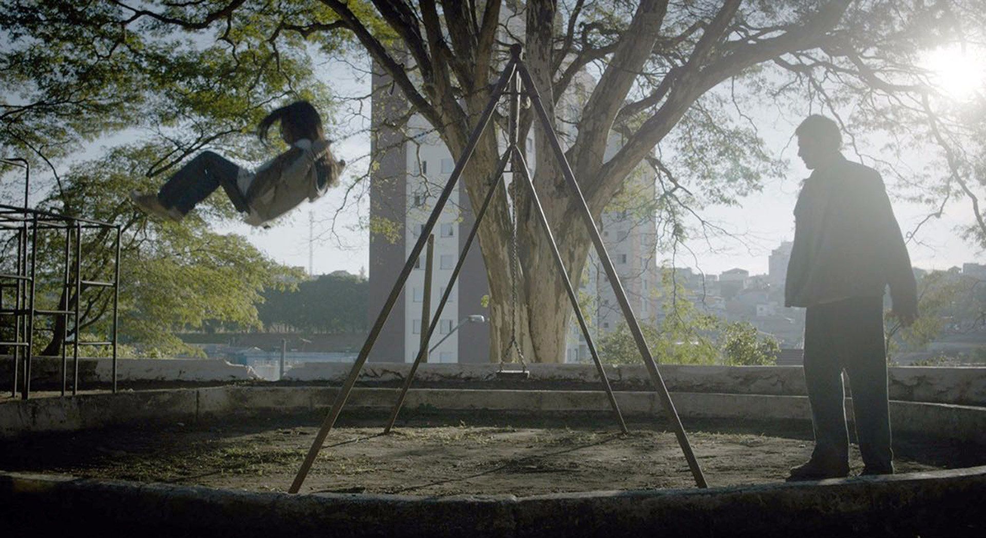 Still do filme 'A sombra do pai' filmado no Butantã A man is standing next to a swing set with a child on it.