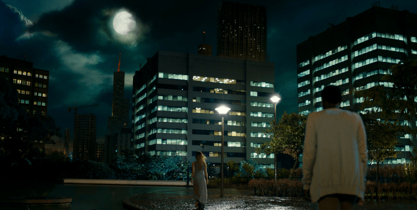 Mulher caminha debaixo de chuva em praça de Alphaville A man and a woman are walking in the rain in front of a city at night.