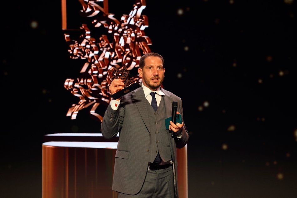Nick Story receives the Sportel Award at Monaco A man in a suit and tie is holding a microphone and a trophy on a stage.