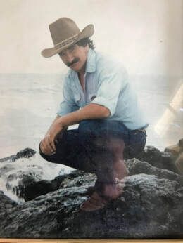 A man wearing a cowboy hat is squatting on a rock near the ocean.