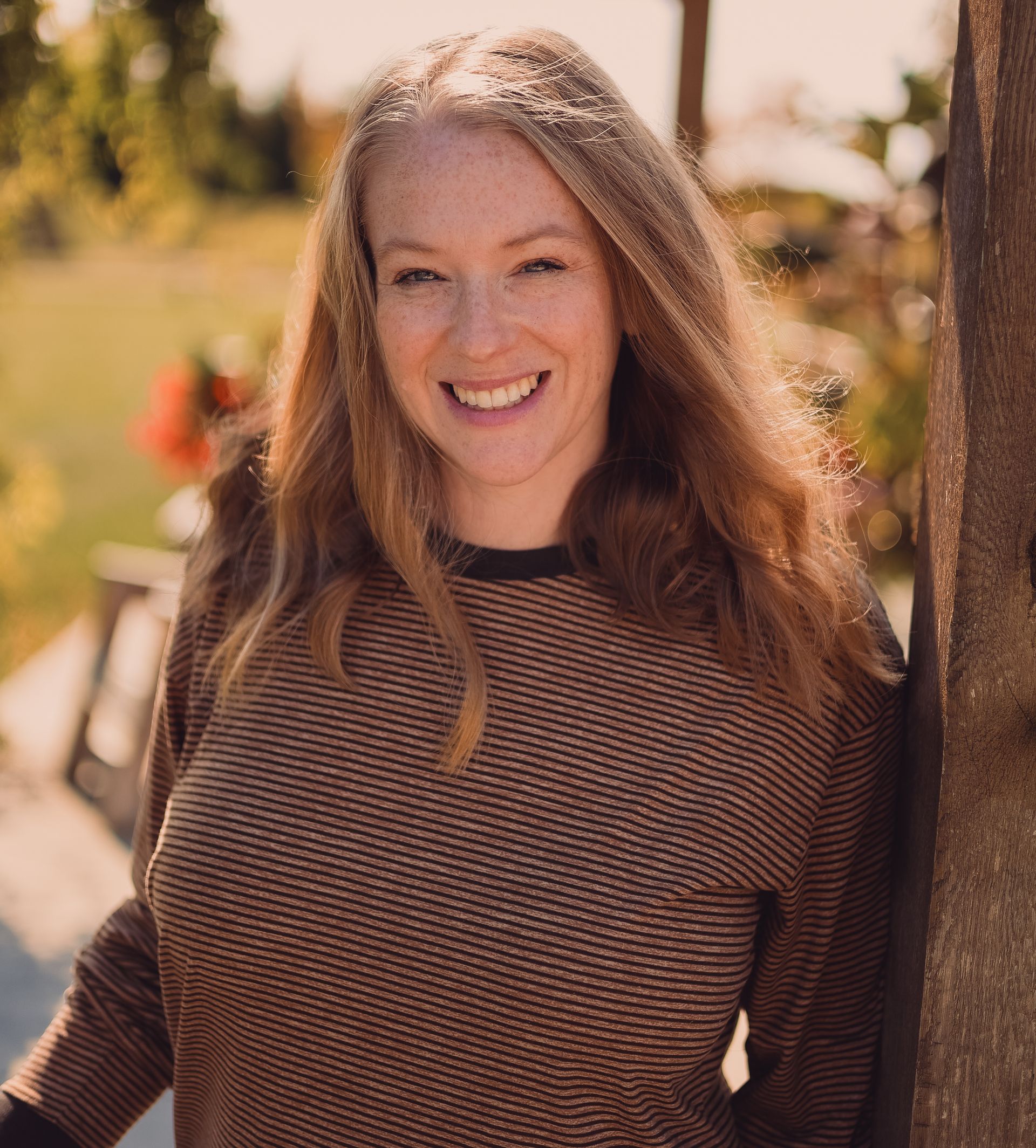Woman with red hair smiles, leaning on a wooden post outdoors, wearing a brown and black patterned sweater.