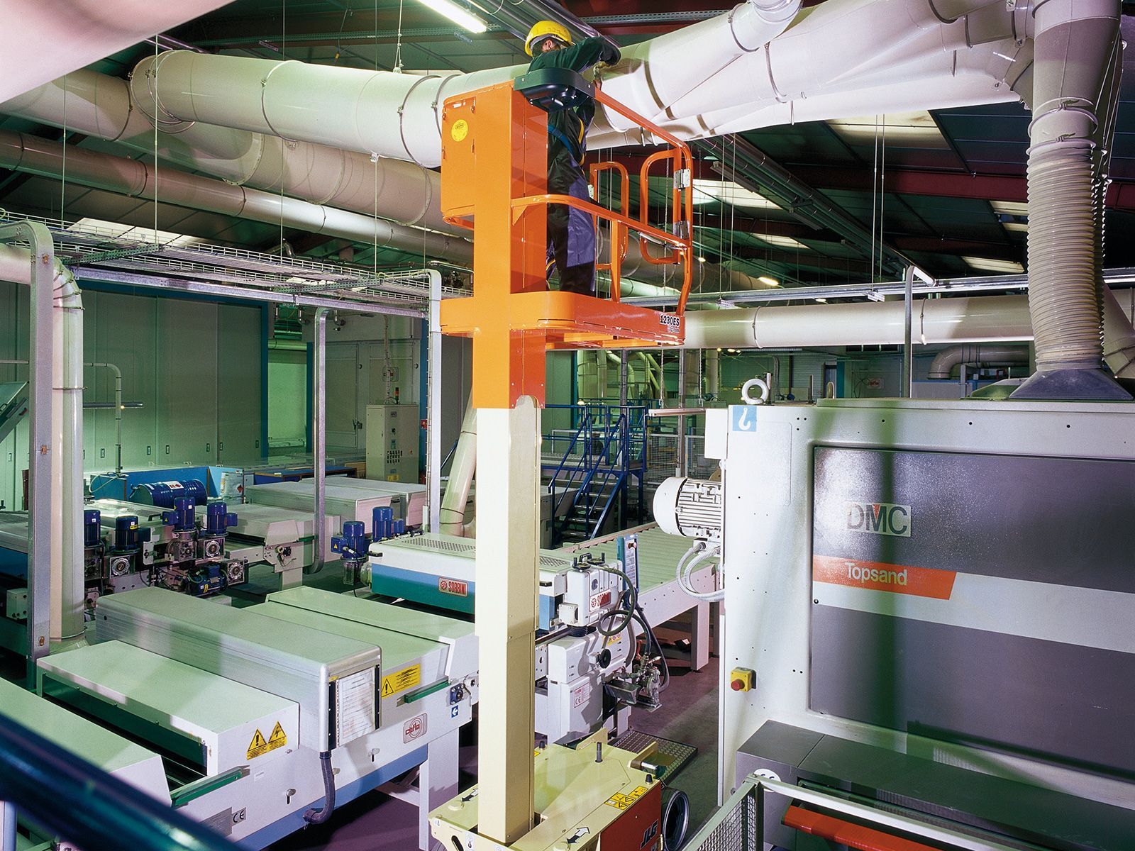 Persona in un ascensore arancione che lavora su condotti sospesi in un ambiente industriale.