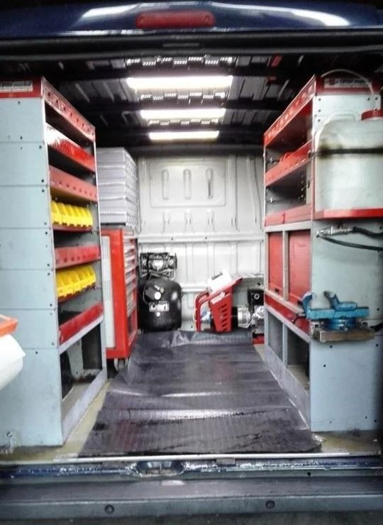Interno di un furgone di servizio con scaffali, utensili e attrezzature disposti lungo le pareti, pavimento nero.
