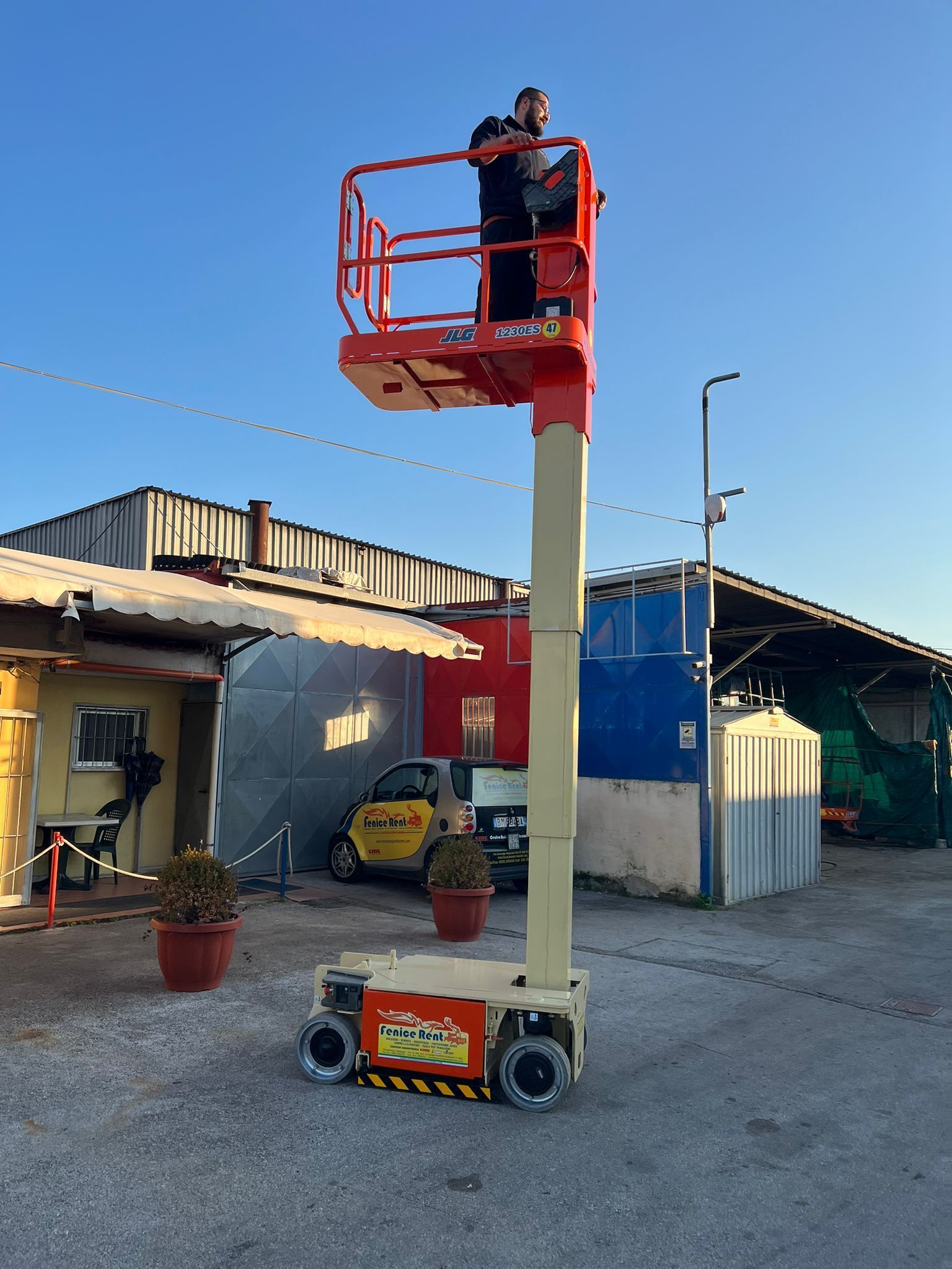 Uomo al lavoro su una piattaforma elevatrice rialzata. Ascensore arancione, cielo azzurro, ambiente industriale.