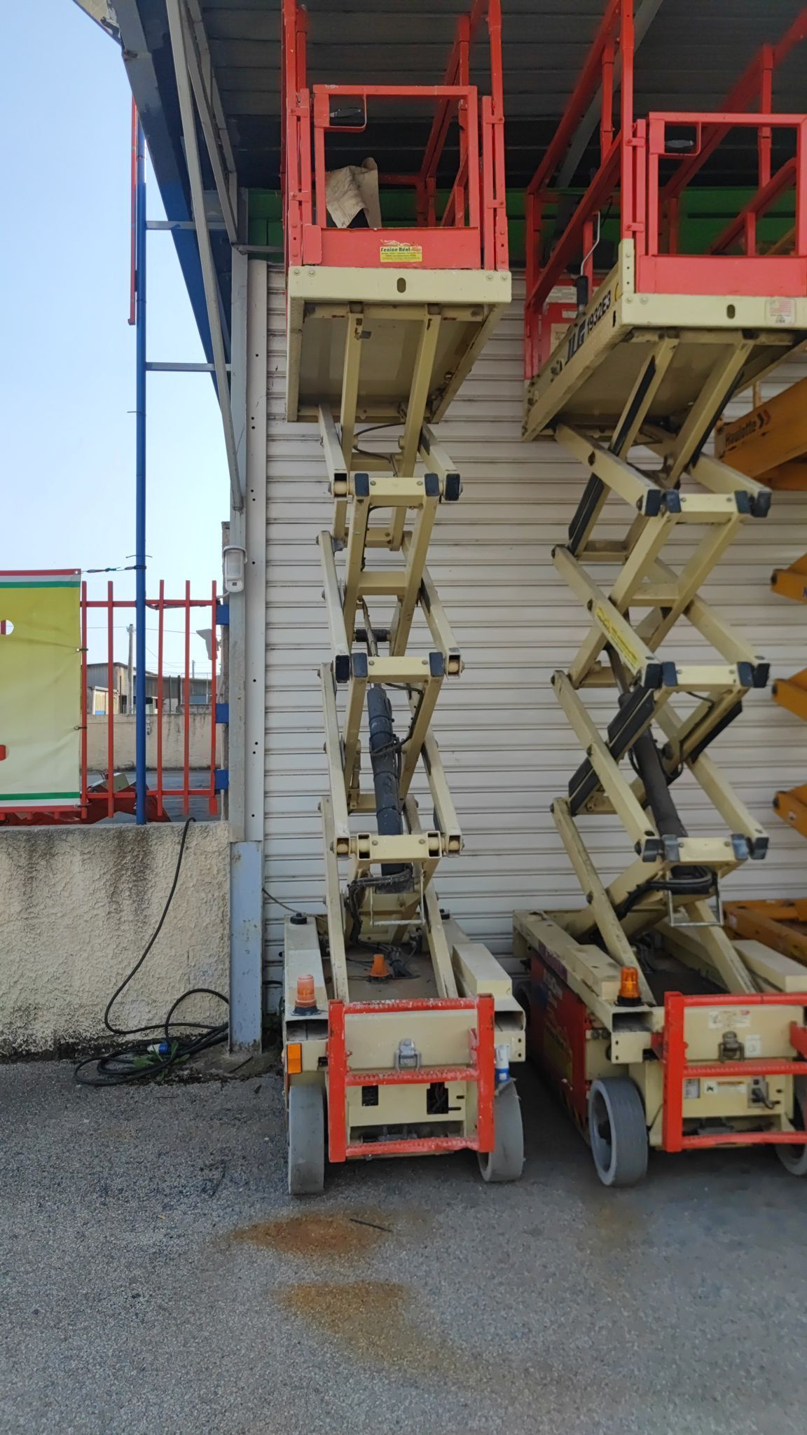 Due piattaforme aeree beige e arancioni parcheggiate contro un muro ondulato. Una è sopraelevata.