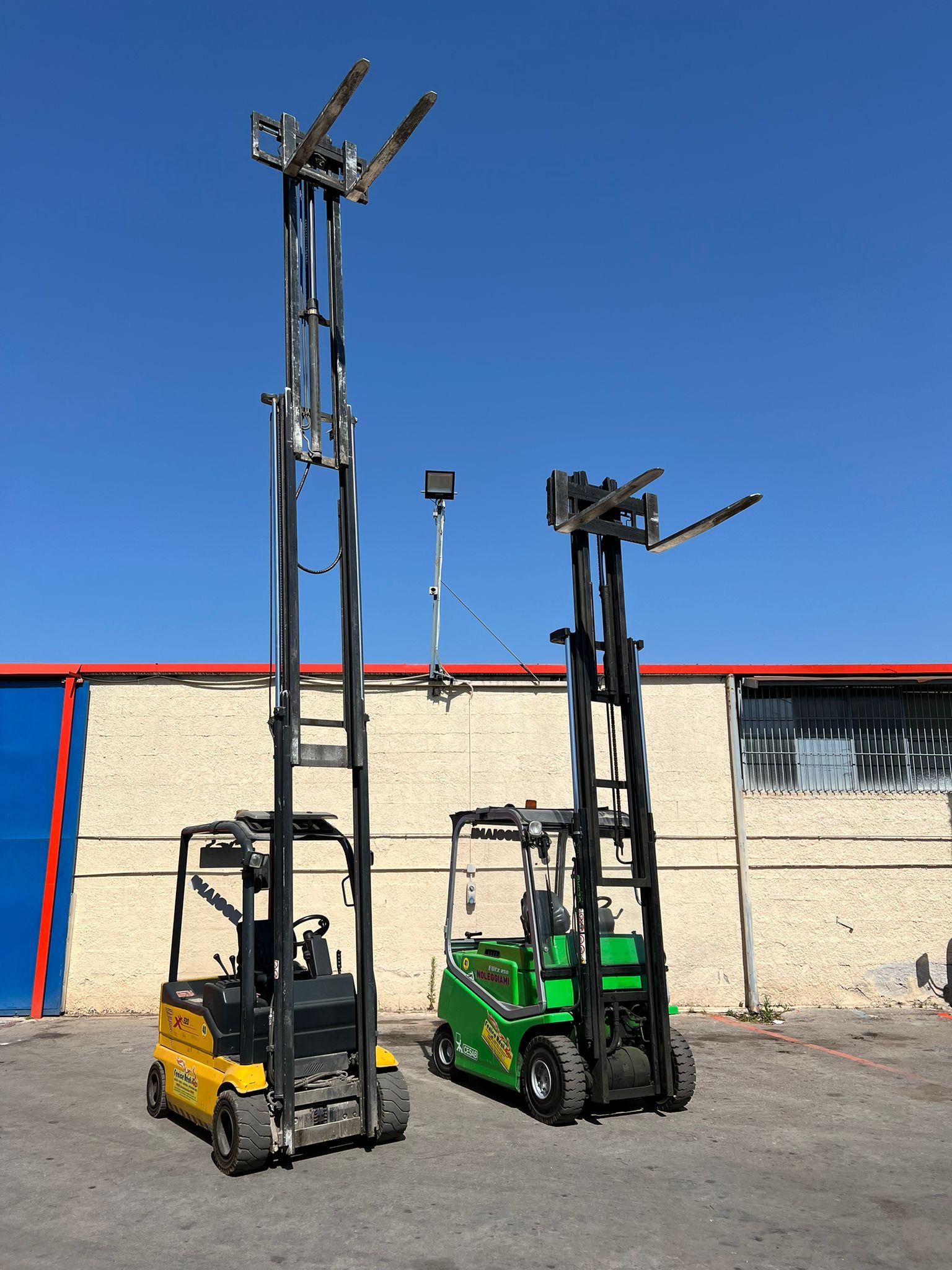 Due alti carrelli elevatori, uno giallo e uno verde, parcheggiati fuori da un edificio contro un cielo azzurro.