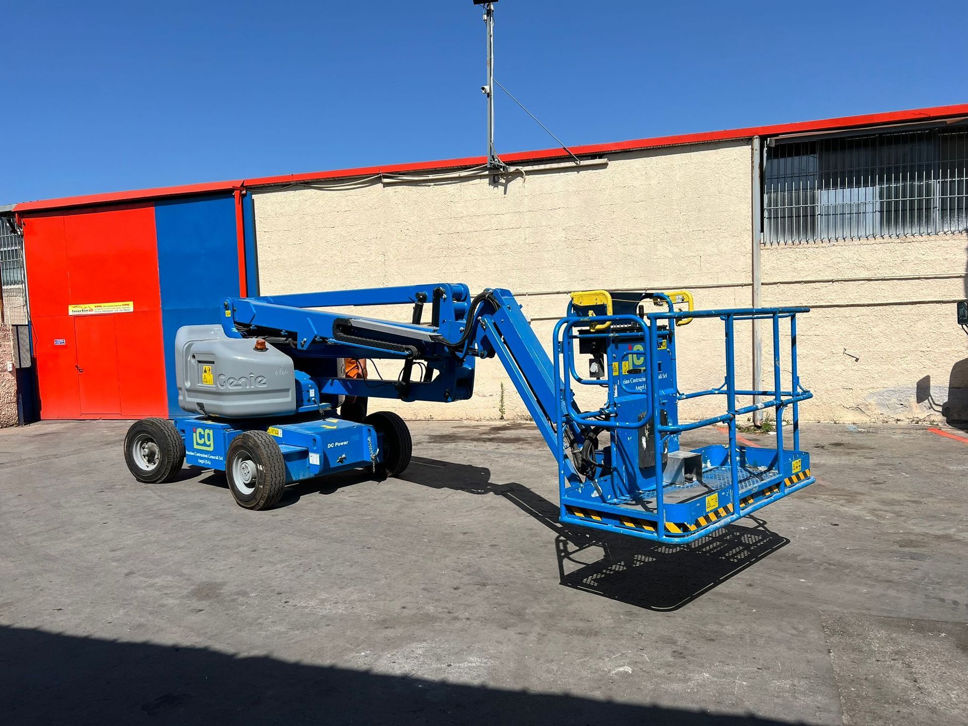 Piattaforma aerea articolata blu parcheggiata su una superficie di cemento; sullo sfondo un edificio rosso e blu.