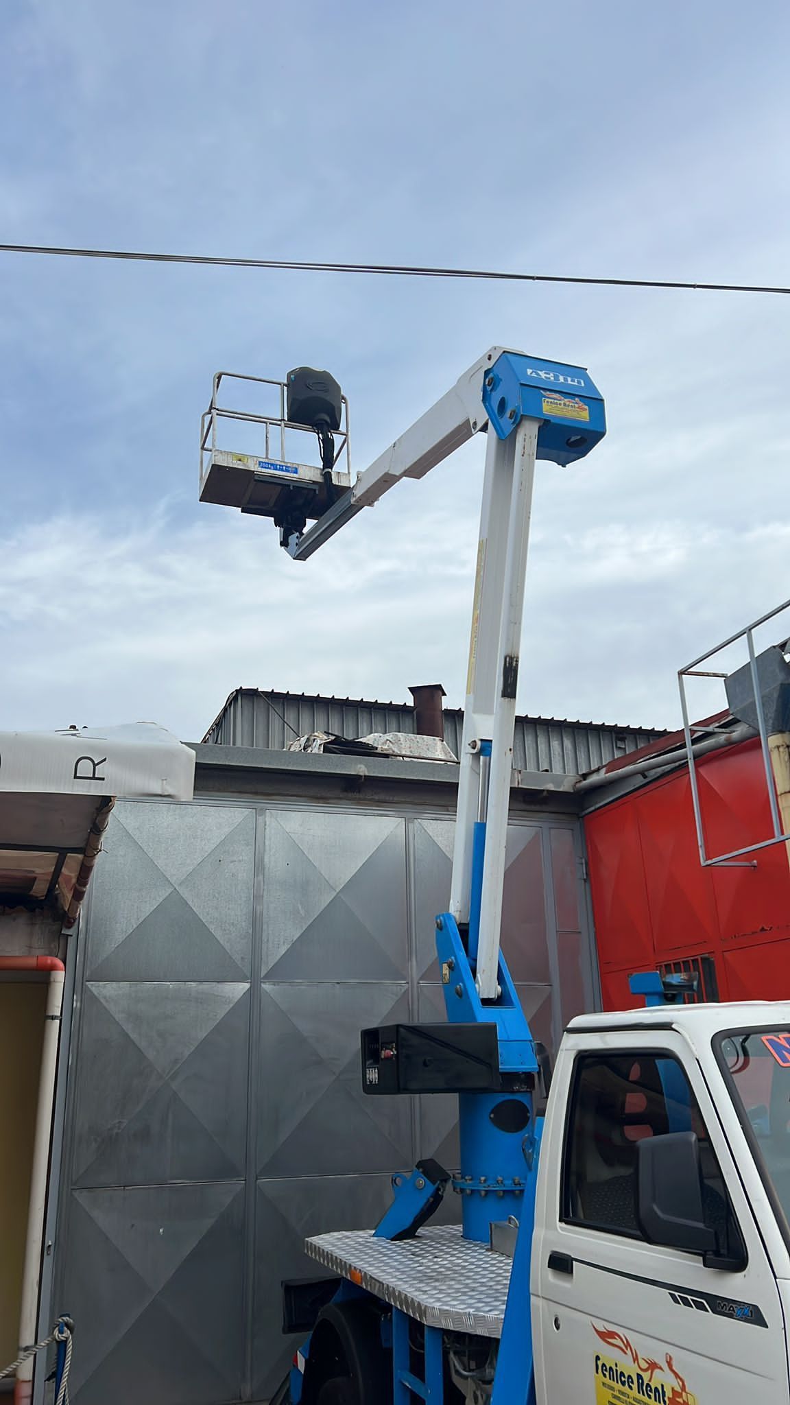 Carrello elevatore aereo blu e bianco con il cestello sollevato contro un cielo nuvoloso.