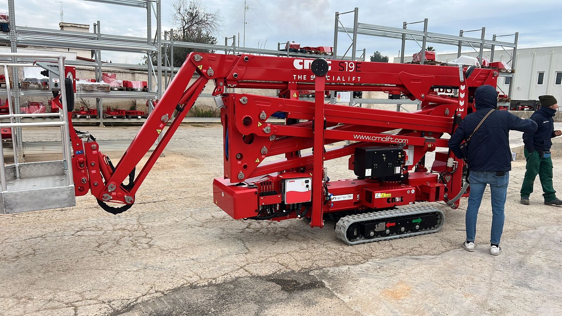 Piattaforma aerea cingolata rossa estesa, con due persone accanto sul marciapiede.