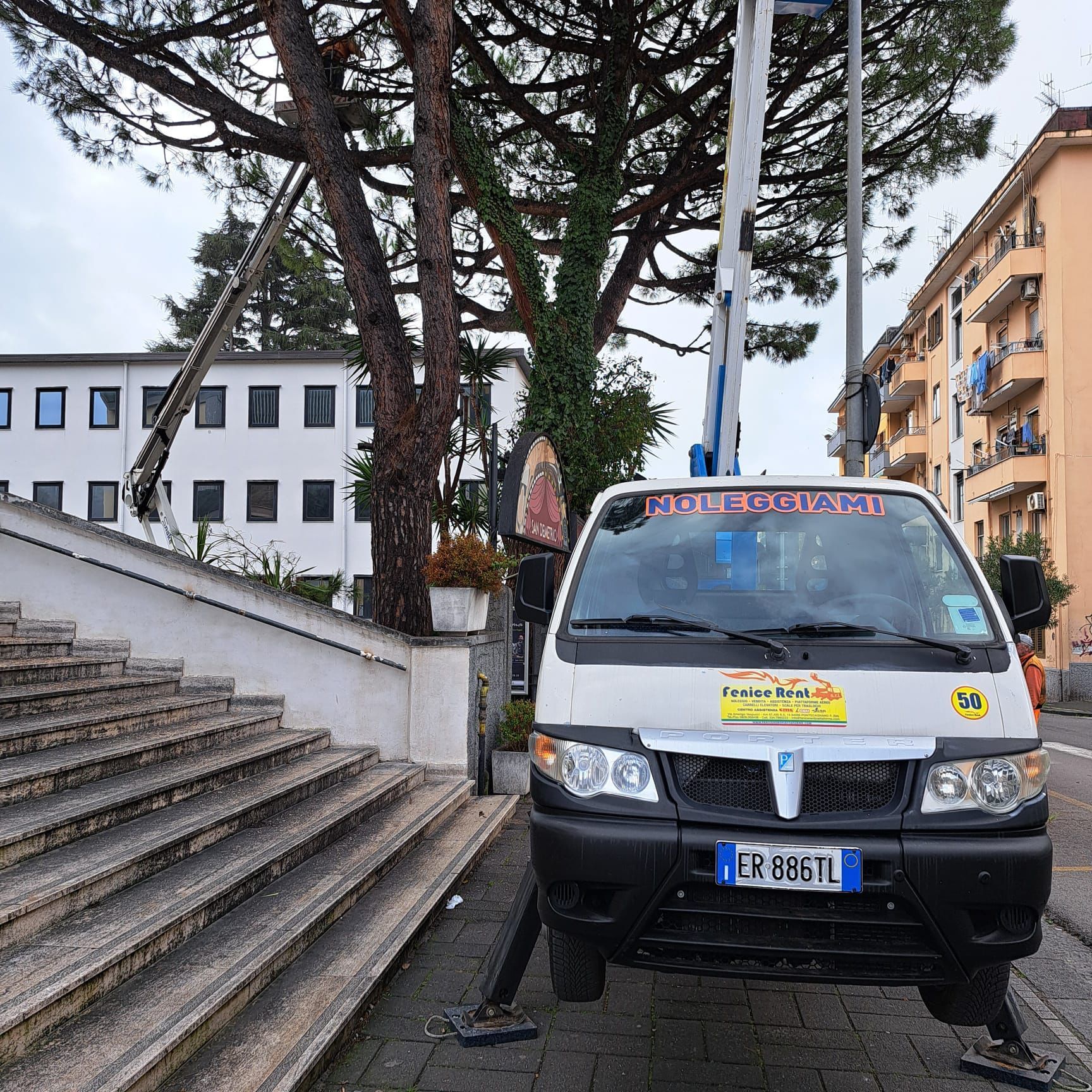 Camion da lavoro bianco con piattaforma di sollevamento rialzata vicino ad alberi ed edifici. Targa ER 688TL.