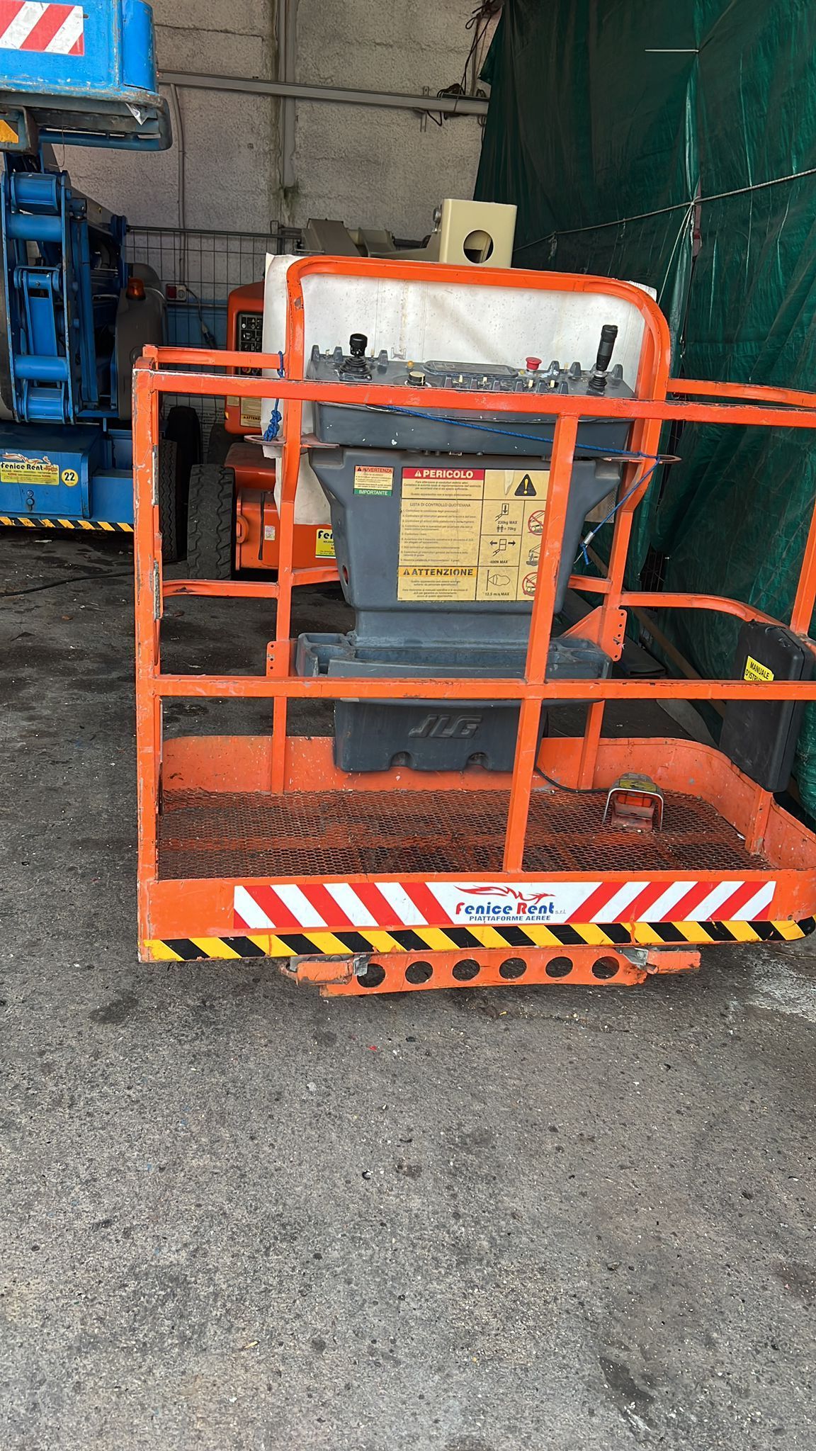 Piattaforma aerea a forbice arancione, pannello di controllo visibile. Ambiente industriale, cemento grigio.