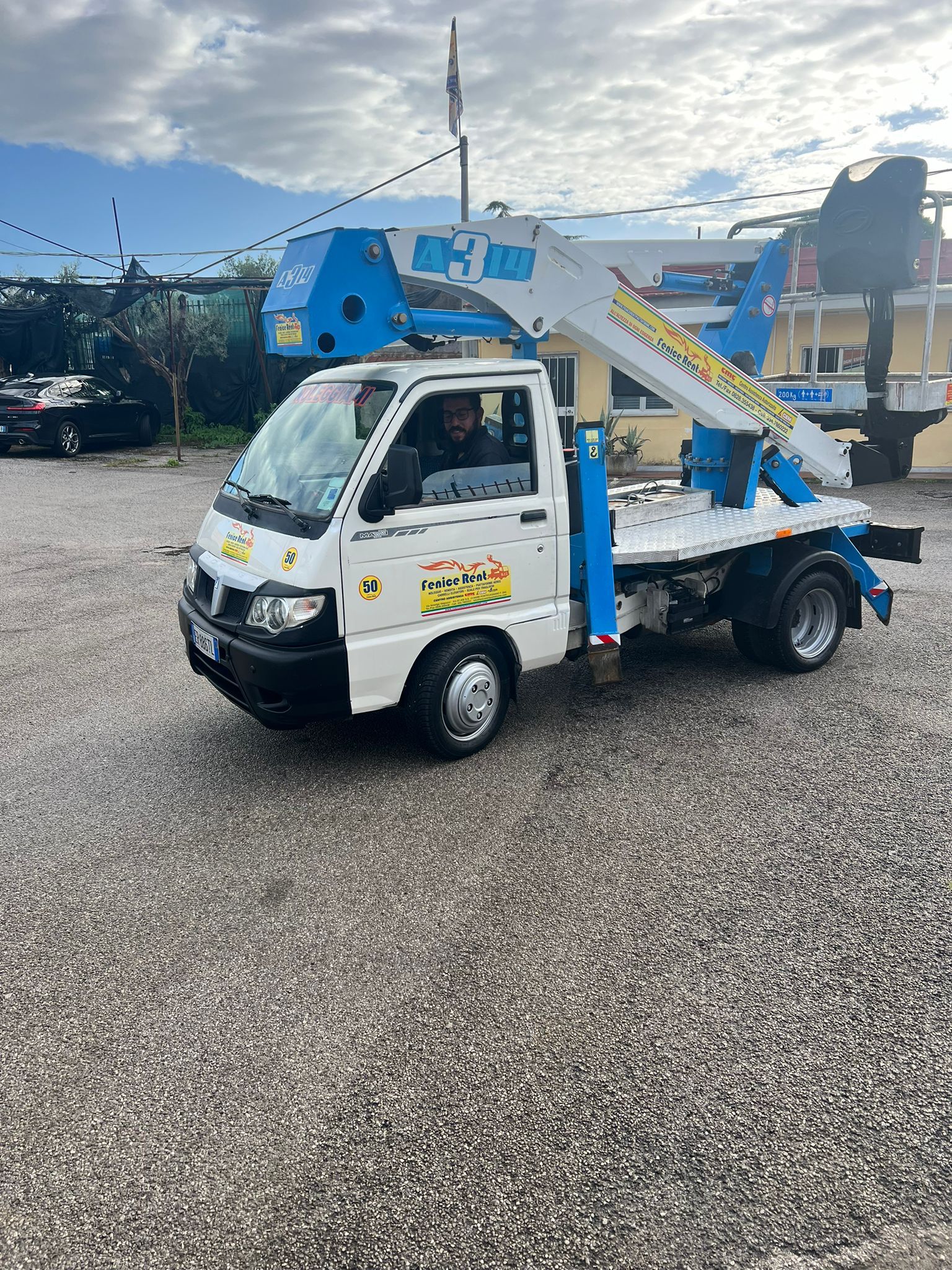 Camion bianco con piattaforma aerea, parcheggiato sulla ghiaia. Braccio di sollevamento blu esteso; conducente all'interno.