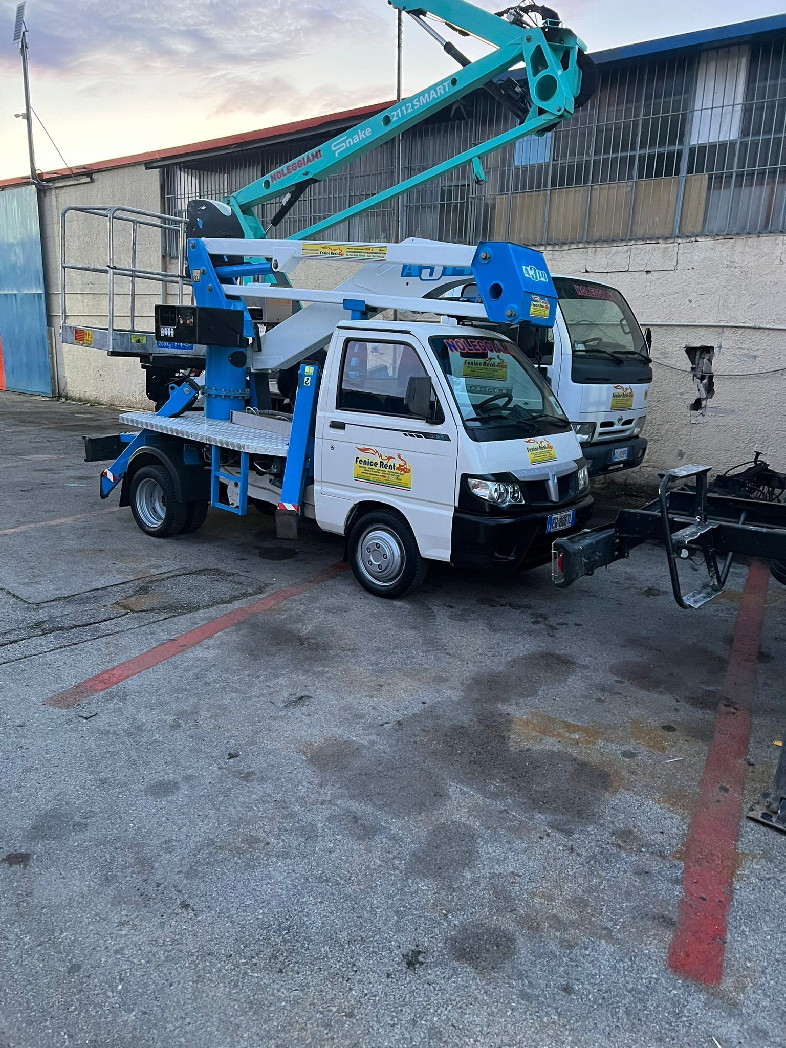 Un camion sollevatore bianco e blu parcheggiato all'esterno. Un secondo camion bianco è parzialmente visibile sullo sfondo.