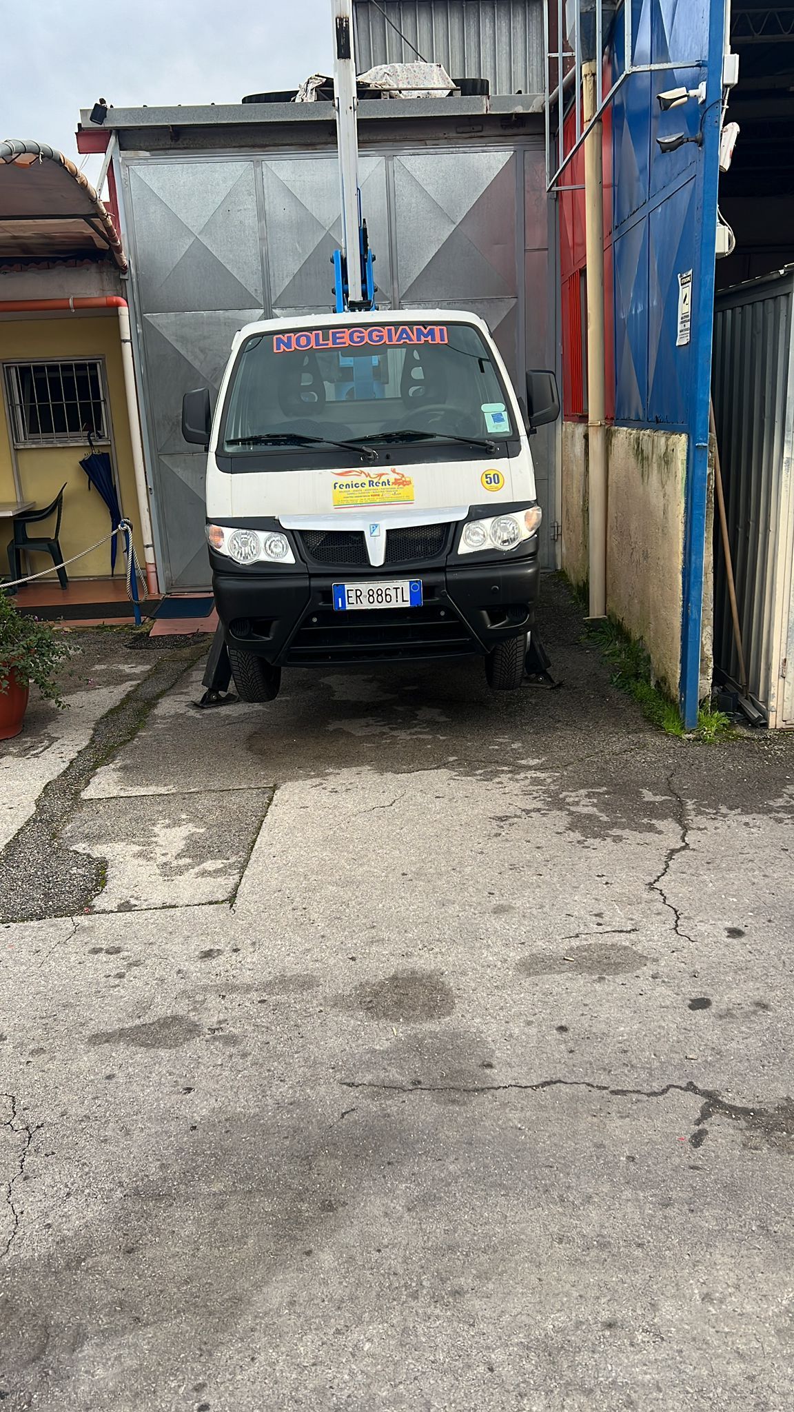 Un camioncino Piaggio Porter bianco parcheggiato in un vicolo, rivolto verso la telecamera, con una grande struttura metallica alle spalle.