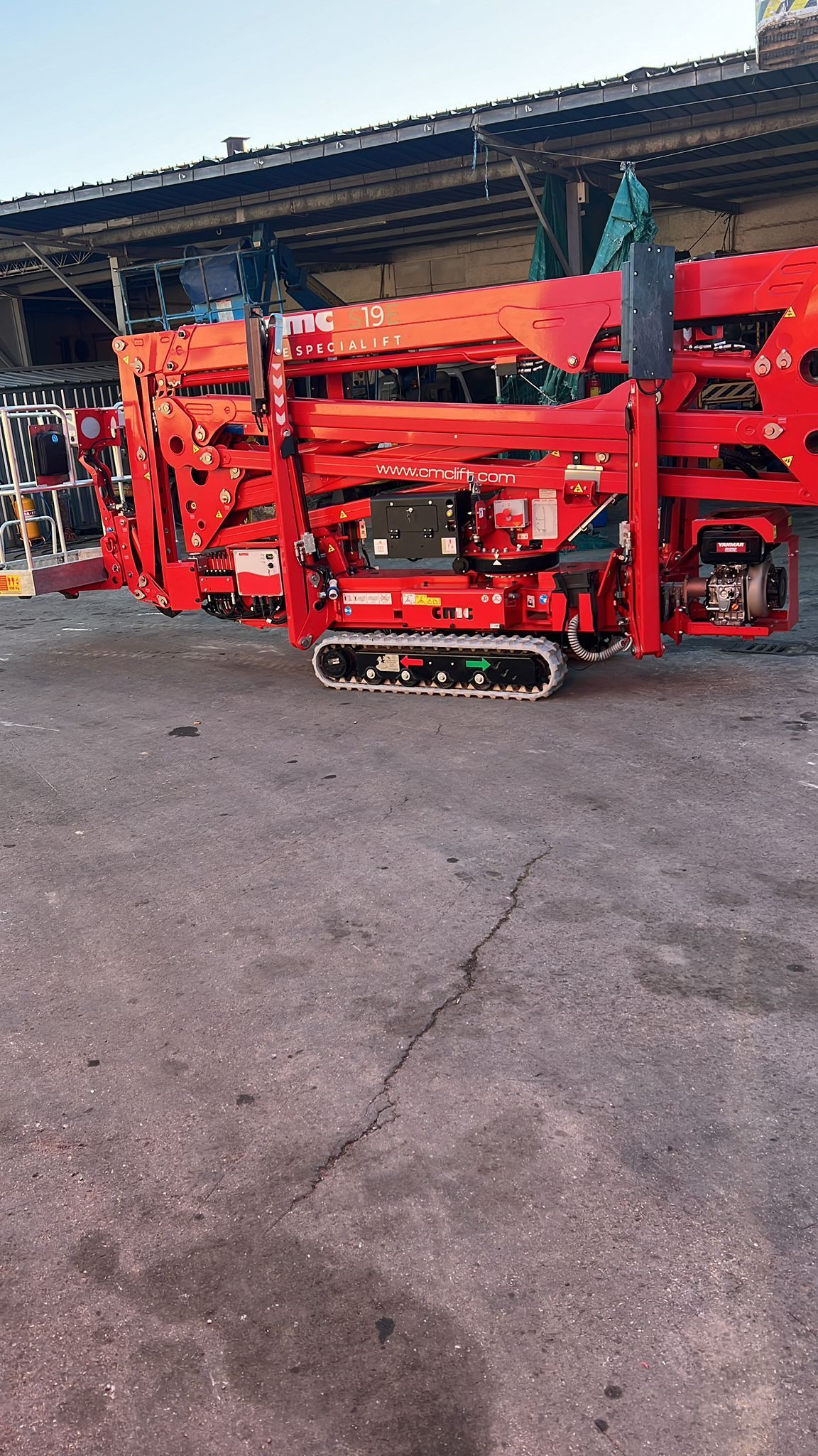 Piattaforma aerea cingolata rossa su asfalto, con un edificio e il cielo sullo sfondo.