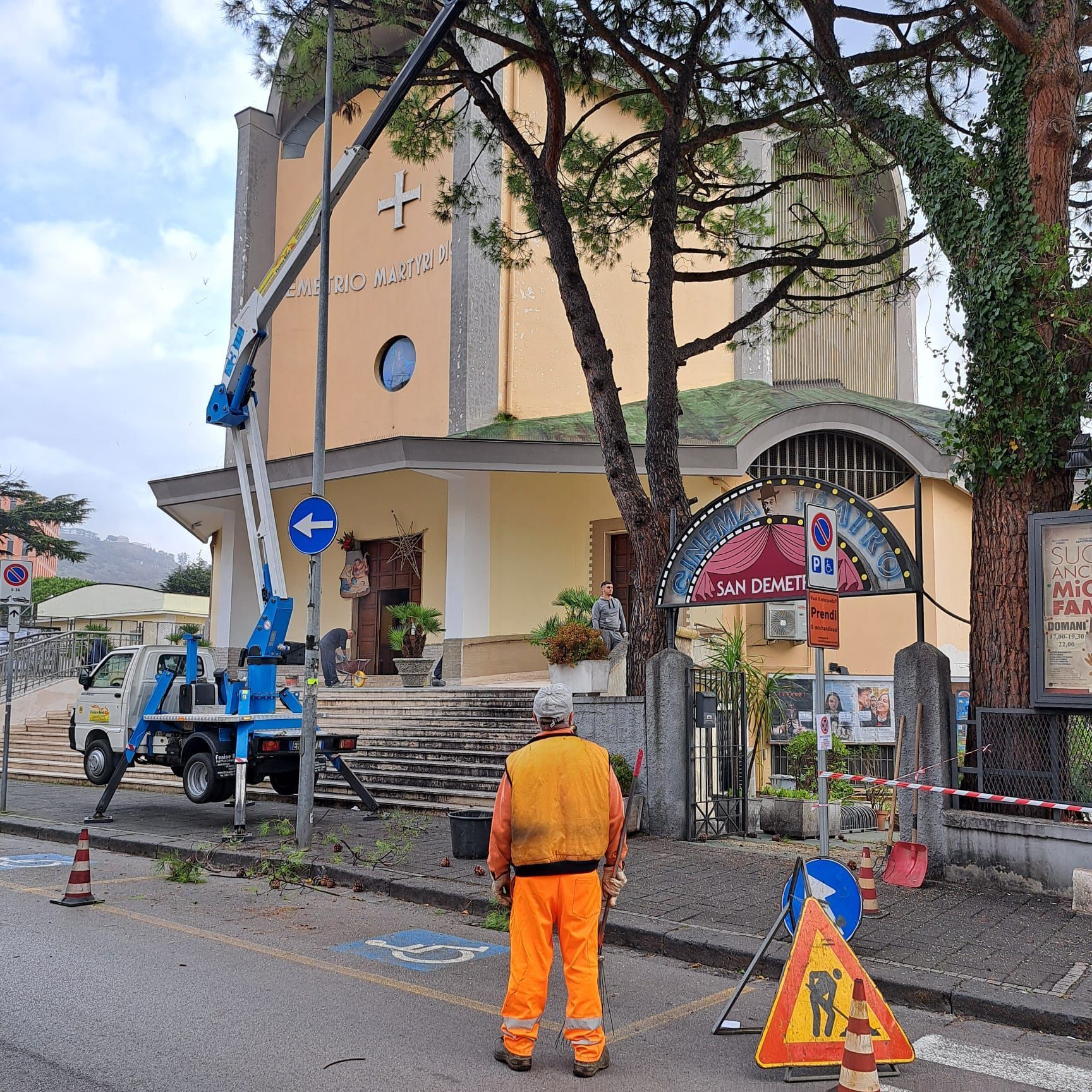 Un uomo con il gilet arancione osserva la potatura degli alberi vicino a una chiesa con un carrello elevatore e cartelli stradali.
