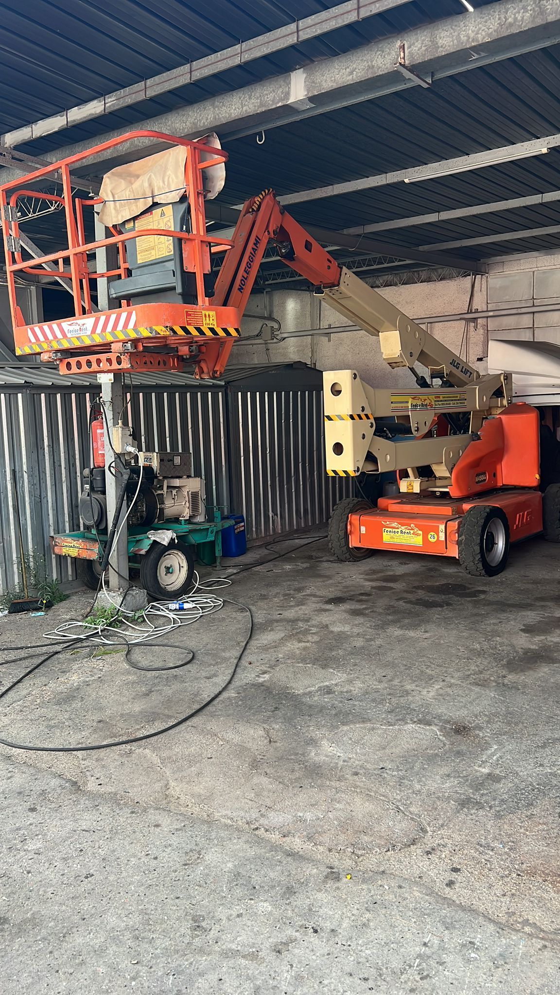 Piattaforma aerea arancione estensibile, con cestello vicino a una parete di lamiera ondulata, all'interno di un edificio.