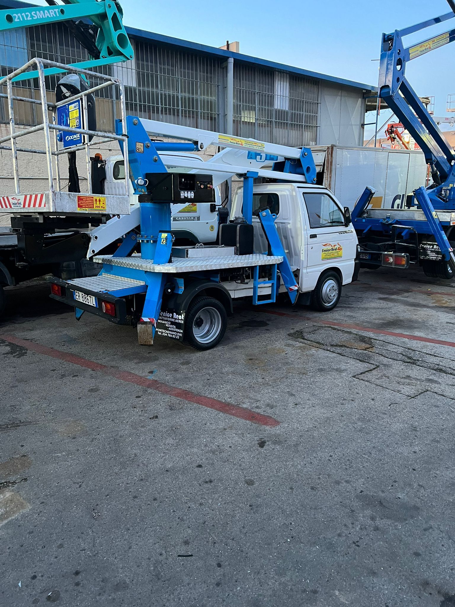 Camion bianco con sollevatore blu, parcheggiato sull'asfalto. Altri sollevatori e un edificio sullo sfondo.