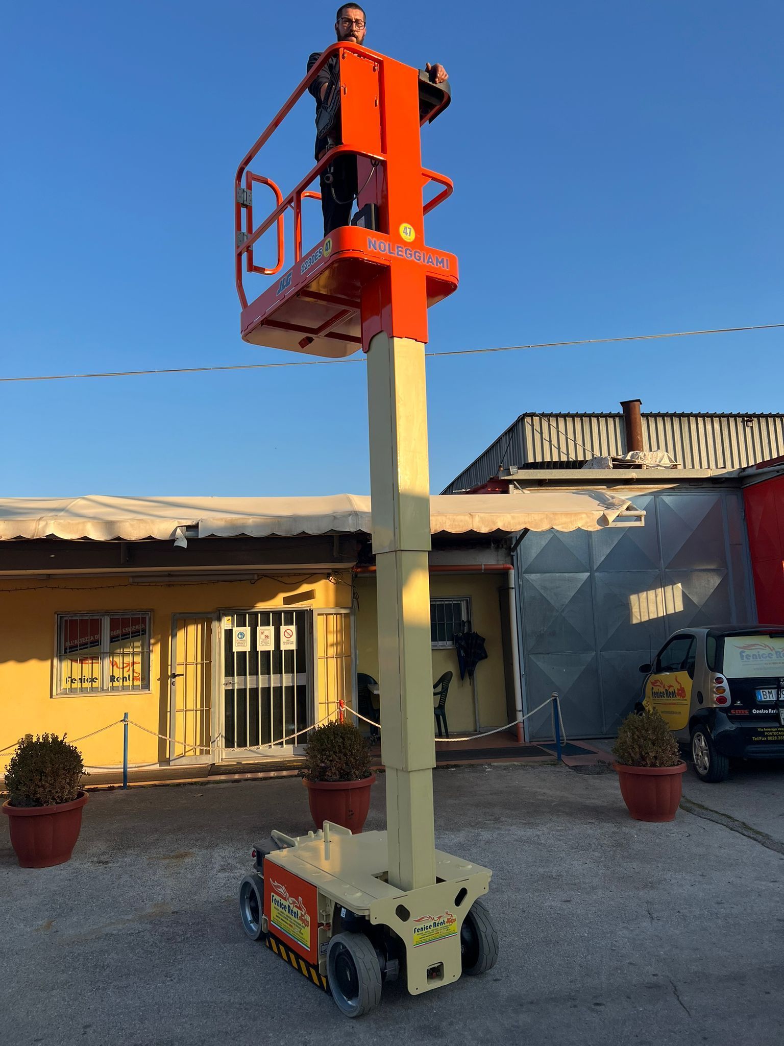 Uomo che manovra una piattaforma aerea arancione e beige. All'aperto, accanto a un edificio, sotto un cielo azzurro.