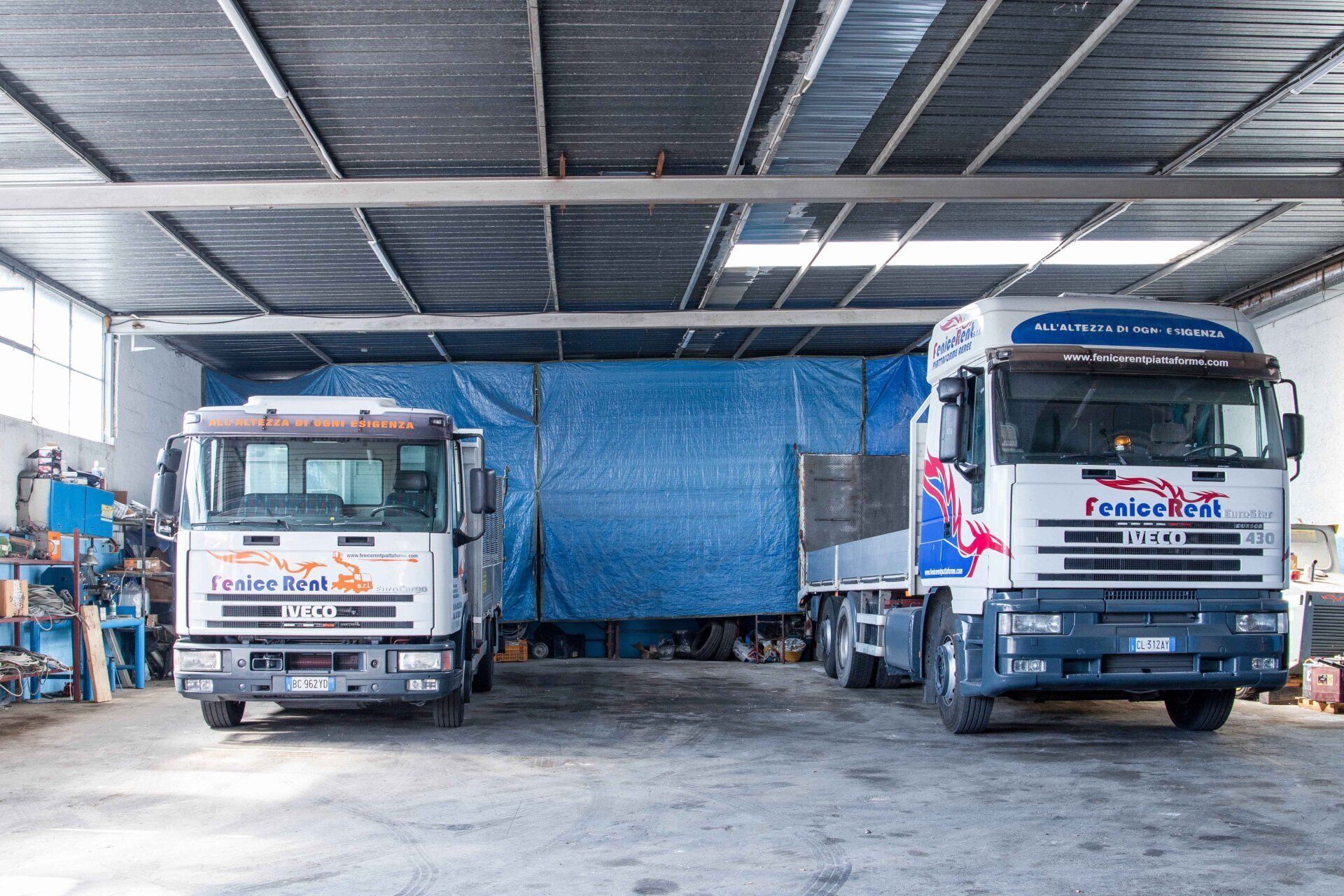 Due camion bianchi parcheggiati all'interno di un'officina con uno sfondo di teloni blu.