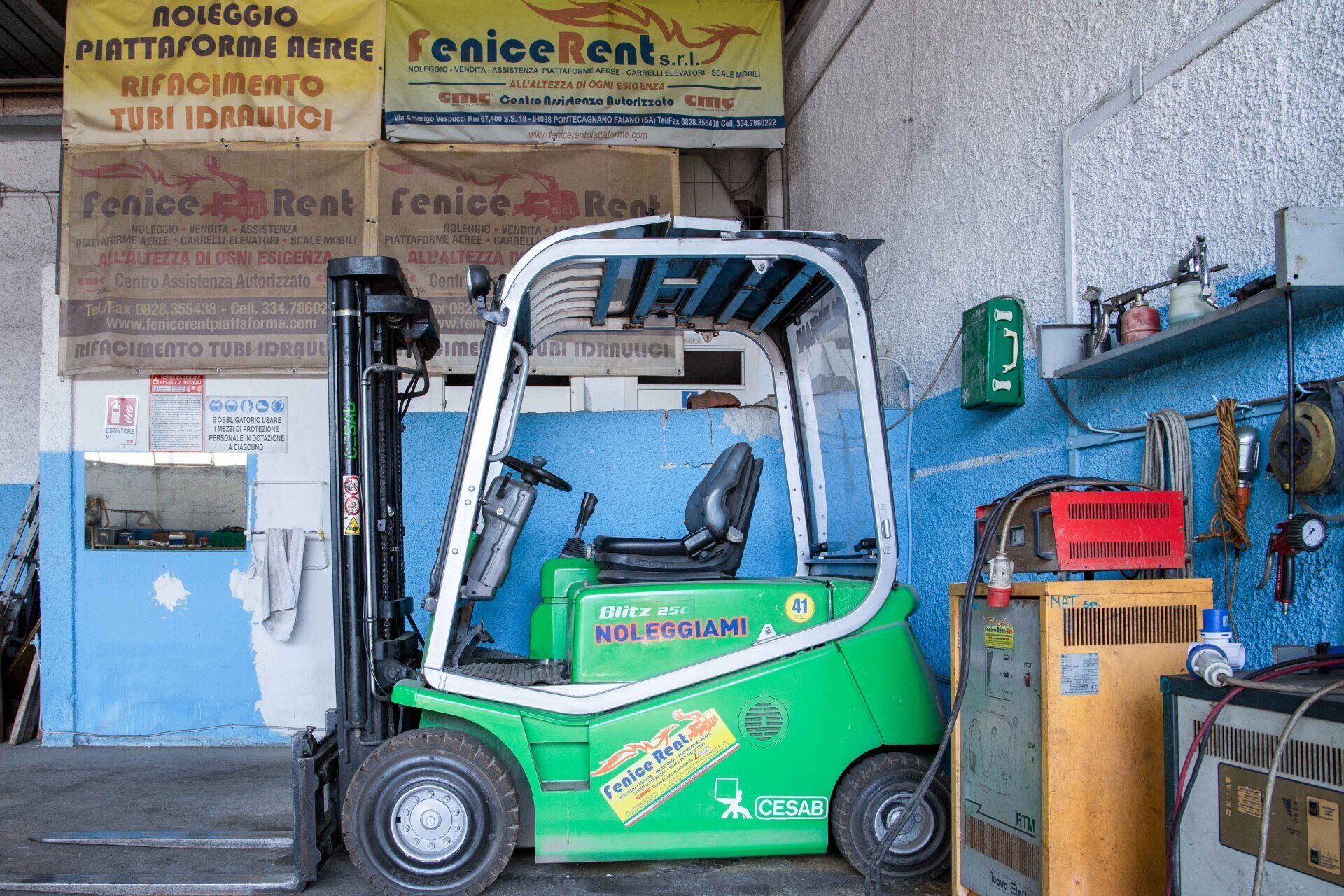 Carrello elevatore verde all'interno di un'officina con attrezzi e segnaletica.