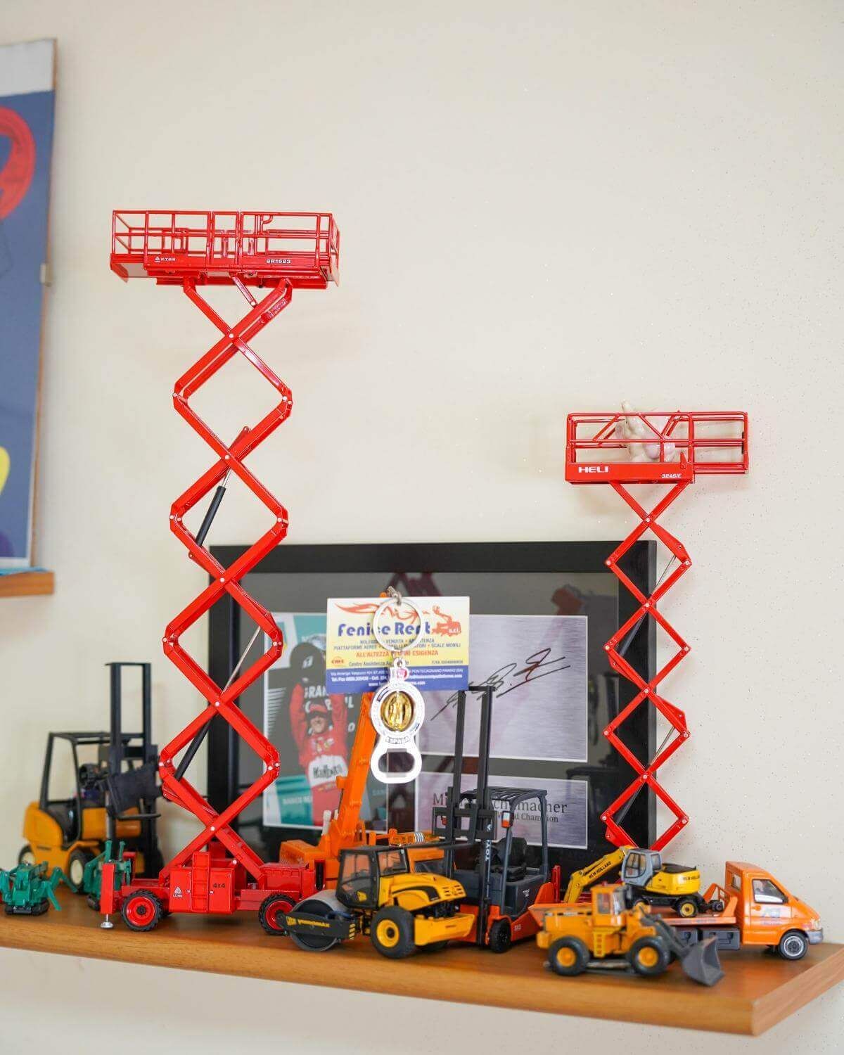 Scaffale di veicoli da costruzione giocattolo, tra cui piattaforme aeree rosse, su una parete bianca.