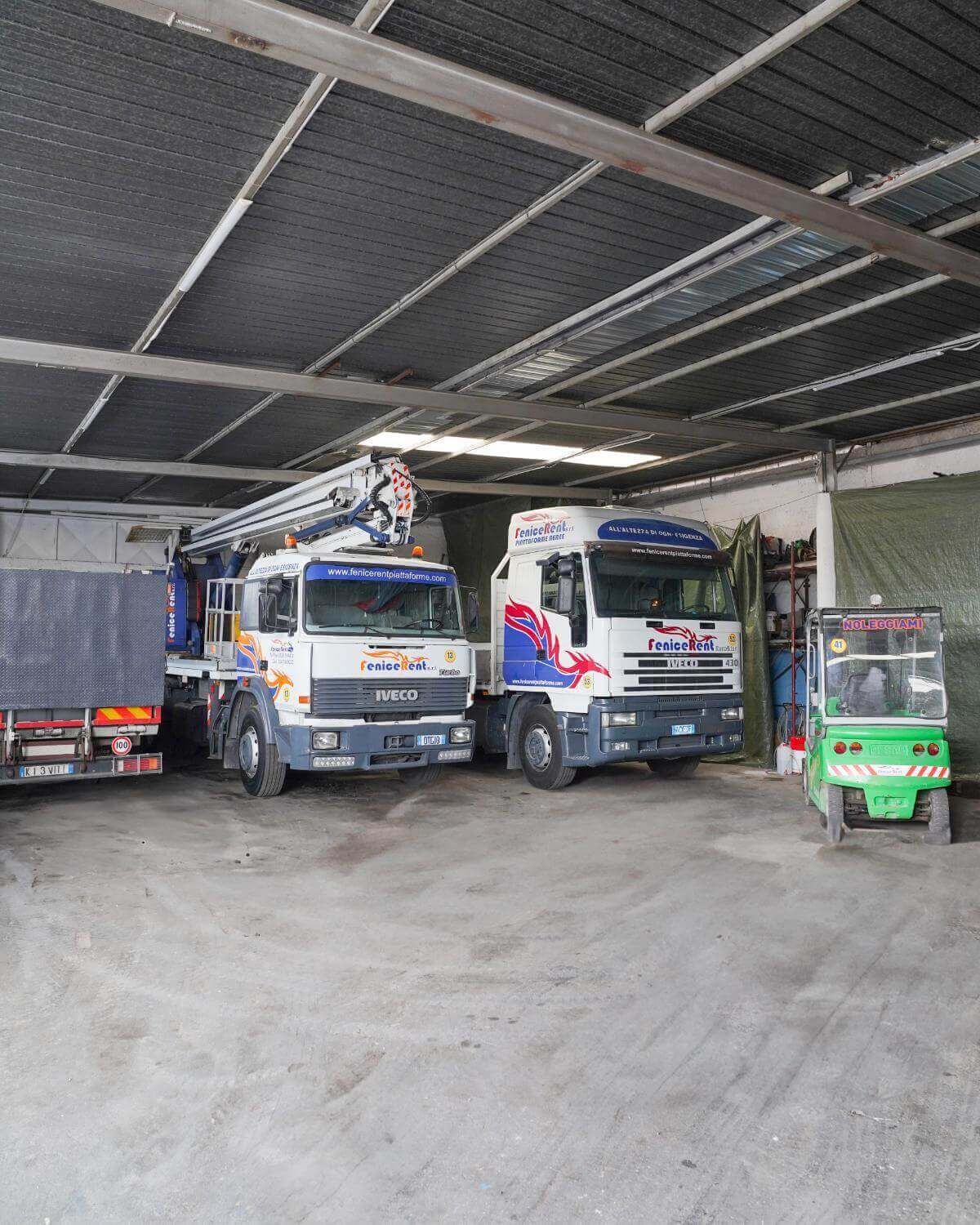 Camion parcheggiati all'interno di un magazzino. Include un carrello elevatore, camion bianchi e blu, pavimento in cemento e soffitto in metallo.