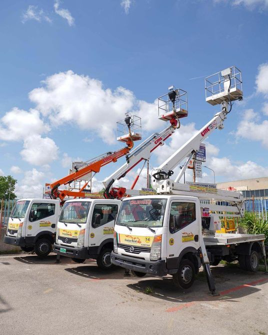 Tre camion bianchi con piattaforme aeree in un parcheggio sotto un cielo azzurro.
